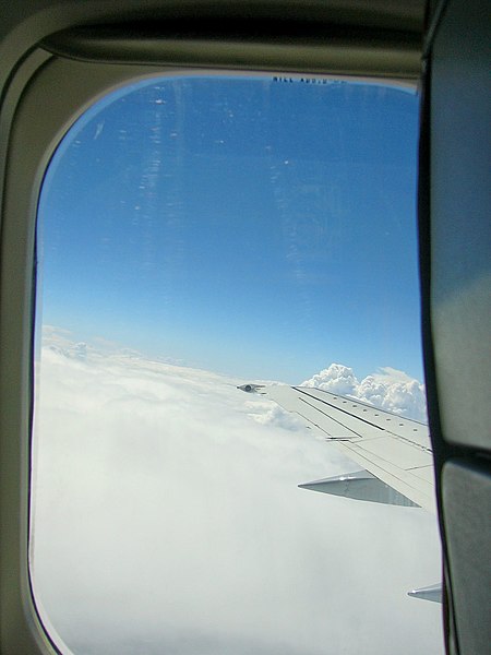 Vista dall’interno di un volo di linea, con l’oblo in vetroplastica che mostra l’ala sinistra di un aereo sorvolante un cielo azzurro con nuvole bianche.