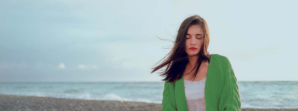 Donna pensierosa in riva al mare con maglione verde, vento tra i capelli e cielo azzurro, atmosfera rilassante.