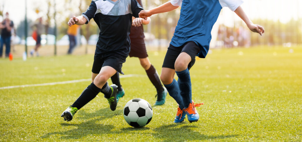 Azione dinamica di calcio su campo in erba sintetica, con due giocatori di squadre avversarie pronti a entrare in un tackle a centrocampo, catturati dai busti in giù durante un momento decisivo di gioco.